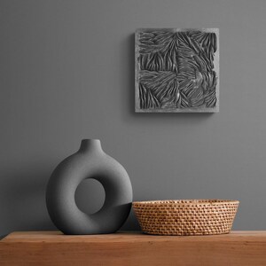 May include: A gray ceramic vase with a donut-shaped opening sits on a wooden table next to a woven wicker basket. A gray square wall decor with a textured pattern hangs on the wall behind the vase.