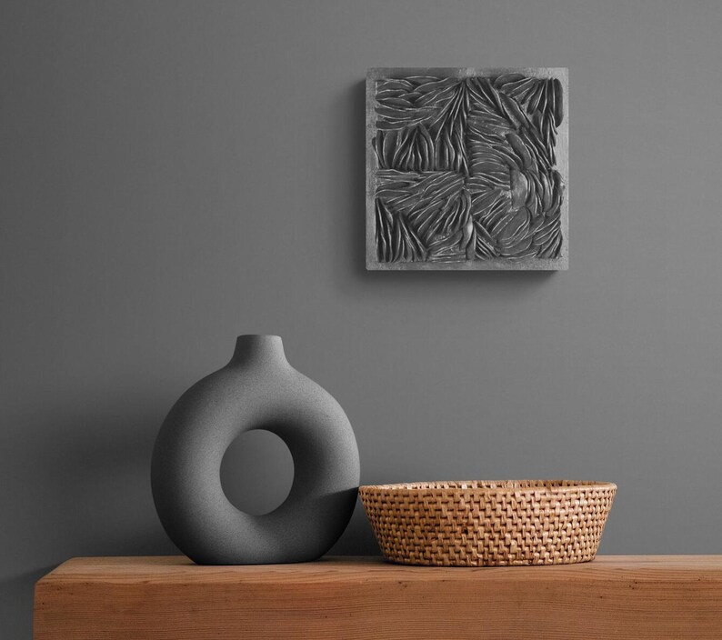 May include: A gray ceramic vase with a donut-shaped opening sits on a wooden table next to a woven wicker basket. A gray square wall decor with a textured pattern hangs on the wall behind the vase.