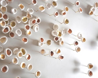 May include: A wall installation featuring numerous small, white, bowl-shaped ceramic elements. Each piece is mounted on a thin metal pin and features a metallic interior finish in shades of gold or copper, creating a textured, repeating pattern.