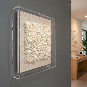 A dimensional art piece, composed of numerous small, off-white, petals arranged densely on a lighter square fabric background, is encased in a clear acrylic box frame. The artwork is mounted on a dark gray wall in what appears to be a modern luxury home entrance. The shot is a side view and a close-up, allowing a clear view of the texture and framing. In the soft-focus background, part of the entrance area is visible, featuring a wooden console table with books and a decorative object.