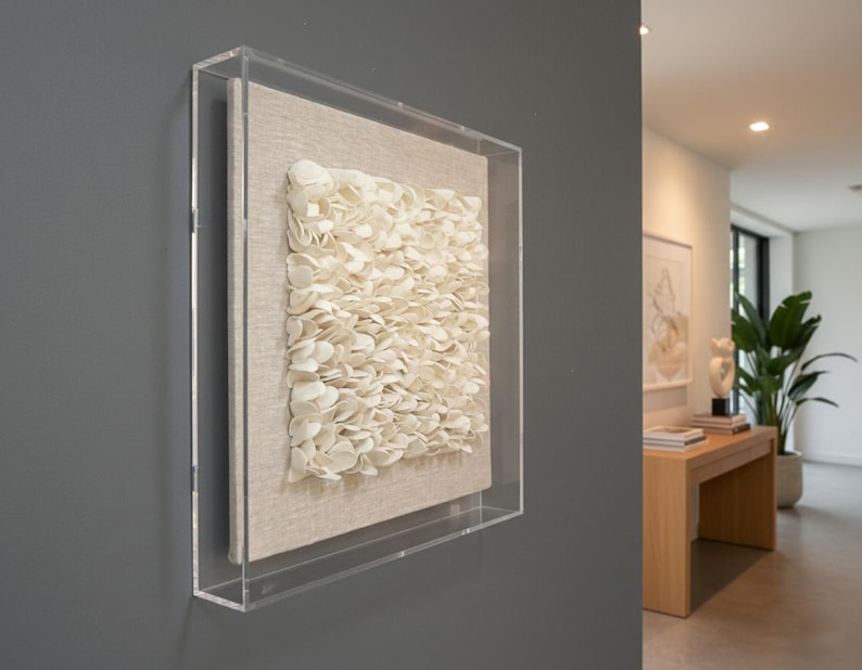 A dimensional art piece, composed of numerous small, off-white, petals arranged densely on a lighter square fabric background, is encased in a clear acrylic box frame. The artwork is mounted on a dark gray wall in what appears to be a modern luxury home entrance. The shot is a side view and a close-up, allowing a clear view of the texture and framing. In the soft-focus background, part of the entrance area is visible, featuring a wooden console table with books and a decorative object.