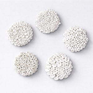 Op de afbeelding: Vijf witte, cirkelvormige keramische objecten met een textuur, gerangschikt in een stervormig patroon tegen een witte achtergrond. Elk stuk heeft een unieke opstelling van kleine, afgeronde vormen, wat een tactiele uitstraling creëert.