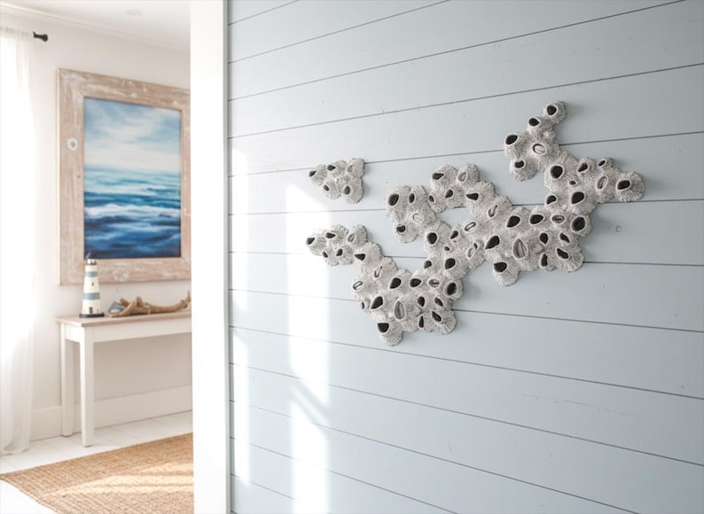 A wall installation made from multiple barnacle-like sculptures is displayed on a light blue wooden slat wall in a bright coastal-themed room. The individual sculptures, each roughly 26x18cm, are white with dark openings, arranged to connect and repeat in various directions, forming a larger organic shape. In the background, a light-filled doorway reveals a glimpse of another room with a framed seascape painting, a white console table, a small lighthouse decoration, and a woven rug.