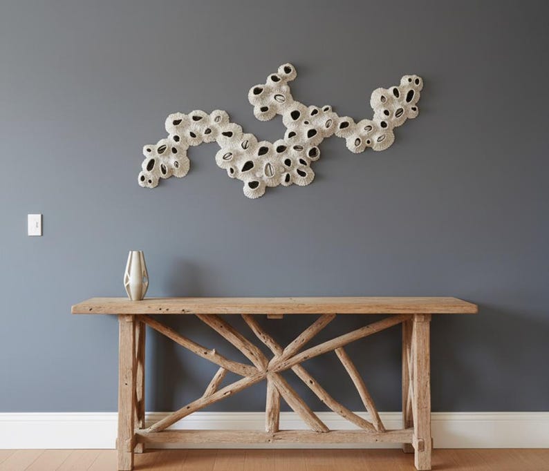 A stylish entryway features a rustic driftwood console table topped with a sleek silver vase. Above the table, a unique white coral-like sculpture, made of individual pieces arranged in an organic, flowing pattern, adorns the slate grey wall. The overall aesthetic is natural and contemporary, with warm wooden flooring extending into the background where a mirror reflects a glimpse of another room.