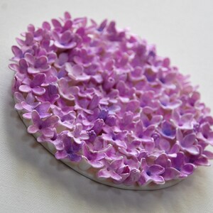 May include: A circular wall art piece made of paper flowers. The flowers are all shades of purple and are arranged in a tight cluster. The piece is mounted on a white background.