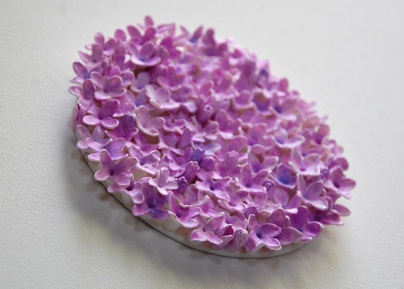 May include: A circular wall art piece made of paper flowers. The flowers are all shades of purple and are arranged in a tight cluster. The piece is mounted on a white background.