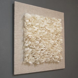 May include: A white wall art piece made of small, organic shapes, resembling petals or leaves, arranged in a textured, layered pattern. The piece is mounted on a beige canvas.