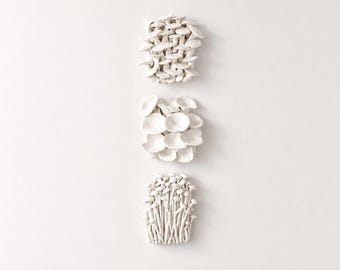 Puede incluir: Tres esculturas de hongos de cerámica blanca dispuestas verticalmente sobre un fondo blanco. La escultura superior se asemeja a un grupo de hongos pequeños apilados. La del medio presenta formas superpuestas en forma de abanico. La inferior tiene tallos largos y delgados.