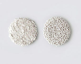 May include: Two white, circular, ceramic objects. One is composed of small, irregular shapes, and the other is made of tiny, spherical beads. Both are set against a plain, white background, creating a minimalist aesthetic. The objects appear to be decorative.