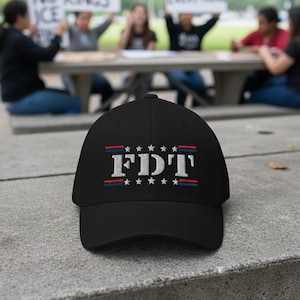 May include: Black baseball cap with the embroidered text "FDT" in white, with red and blue stripes and stars. The cap is sitting on a concrete surface, with people in the background holding signs.