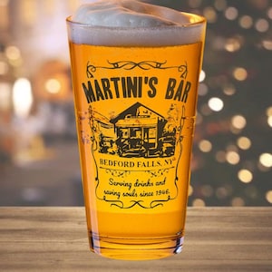 May include: A frosted glass pint glass with a black and white illustration of a building with the text "Martini's Bar, Bedford Falls, NY, Serving drinks and saving souls since 1946."
