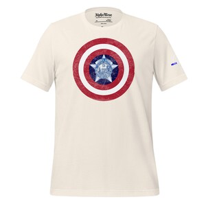 May include: Cream-colored t-shirt featuring a distressed graphic of a red and white shield with a blue star and emblem in the center. The shirt has short sleeves and the text "Alpha Wear" is printed on the collar.