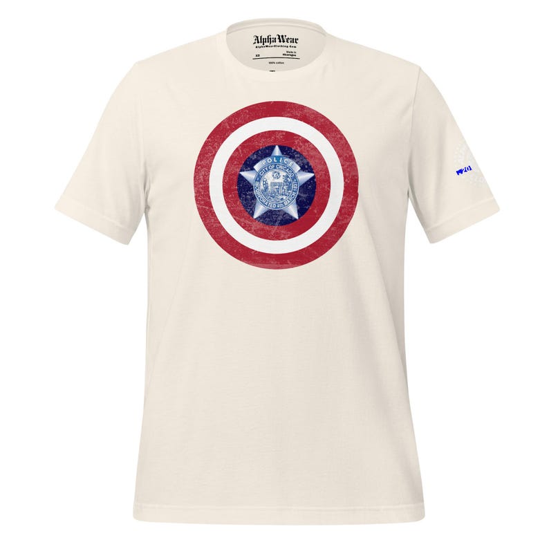 May include: Cream-colored t-shirt featuring a distressed graphic of a red and white shield with a blue star and emblem in the center. The shirt has short sleeves and the text "Alpha Wear" is printed on the collar.