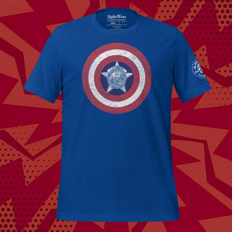 May include: Blue t-shirt featuring a shield design with red, white, and blue concentric circles and a central star emblem. The shirt has short sleeves and a crew neck. The brand name "Alpha Wear" is printed on the tag.