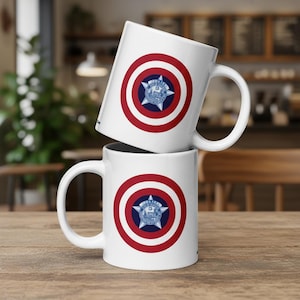 May include: Two white ceramic mugs stacked on a wooden surface. Each mug features a red and white circular design with a blue star emblem in the center. The emblem has the text "POLICE" and a city name.