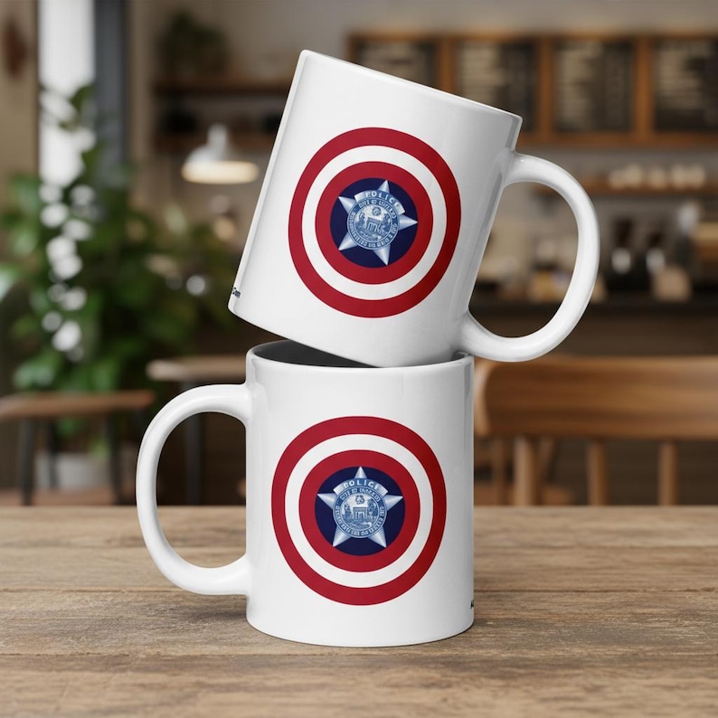 May include: Two white ceramic mugs stacked on a wooden surface. Each mug features a red and white circular design with a blue star emblem in the center. The emblem has the text "POLICE" and a city name.