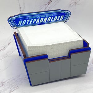 Puede incluir: Un portanotas gris y azul con el texto "NOTEPADHOLDER" y "TOMORROWLAND TRANSIT AUTHORITY" en blanco. El soporte está lleno de una pila de papel de notas blanco. El diseño es de estilo retro.