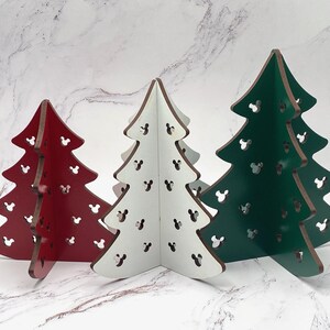 May include: Three wooden Christmas tree decorations in red, white, and green. Each tree features cut-out Mickey Mouse head shapes. The trees are freestanding and have a brown edge.