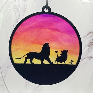 May include: Circular ornament with a black silhouette of a lion, warthog, and meerkat set against a sunset of yellow, orange, and purple. The ornament has a clear border and a small loop for hanging.