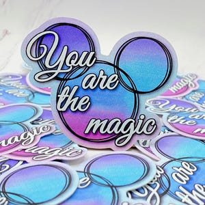 May include: A stack of stickers with a blue, pink, and purple ombre background. The stickers feature a white outline of two overlapping circles and the text "You are the magic" in white.