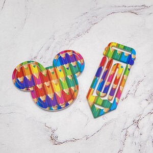 May include: Two colorful wooden cutouts: one in the shape of a mouse head and the other a pencil. Both are decorated with a pattern of colored pencils in various hues. The mouse head cutout is round with two smaller circles for ears. The pencil cutout is a long rectangle with a pointed tip.