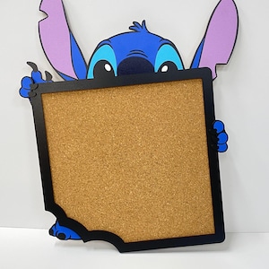 Pode incluir: Um quadro de cortiça azul e preto com o tema Stitch. O rosto e as orelhas do personagem são visíveis acima do quadro, com as mãos segurando a moldura preta. O quadro de cortiça é retangular com um canto faltando.