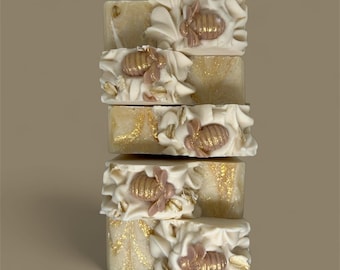 May include: A vertical stack of four rectangular bars of soap. Each bar features a cream-colored base with gold glitter swirls, topped with white textured frosting, embedded oat flakes, and a small, molded bee figure with gold-painted wings.