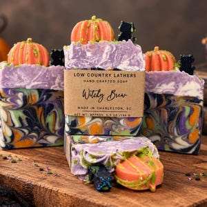 May include: Three rectangular bars of handcrafted soap with a marbled purple, orange, and black swirl pattern. Each bar is topped with a small orange pumpkin and a black cat figure. A brown paper label reads Low Country Lathers Witchy Brew.