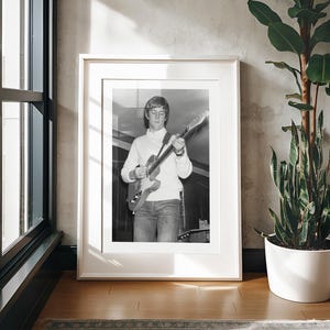 May include: Black and white photo of a young man playing an electric guitar. He is wearing a white turtleneck sweater and blue jeans. The photo is framed in a white frame.