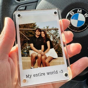 May include: A clear acrylic photo frame with a printed photograph of a couple. The photo is of a man and woman sitting together. The text "My entire world <3" is printed at the bottom of the frame.