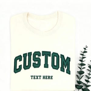 May include: Cream-colored sweatshirt with the word "CUSTOM" arched across the chest in dark green, outlined in a lighter green. Below, the text "TEXT HERE" is also in dark green. The sweatshirt is folded neatly on a white surface.