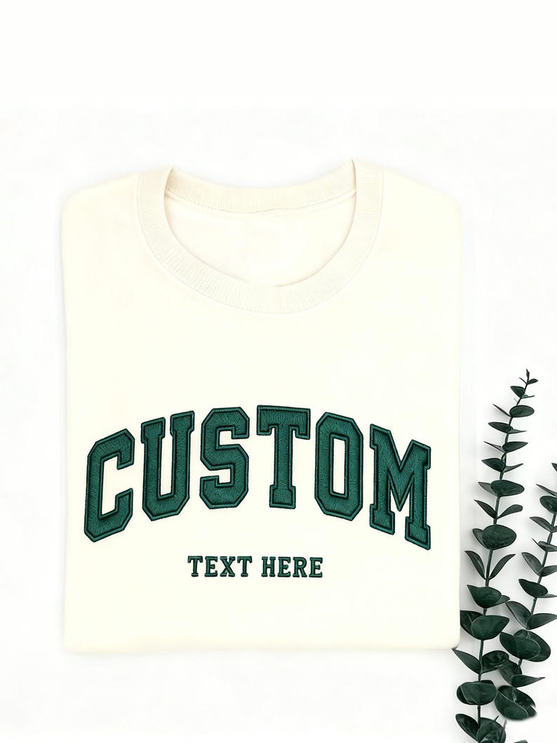 May include: Cream-colored sweatshirt with the word "CUSTOM" arched across the chest in dark green, outlined in a lighter green. Below, the text "TEXT HERE" is also in dark green. The sweatshirt is folded neatly on a white surface.