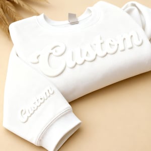 Puede incluir: Una sudadera blanca con la palabra "Custom" en relieve, en fuente cursiva, en el pecho y en la manga izquierda. La sudadera tiene cuello redondo y puños acanalados. El fondo es de color beige neutro.