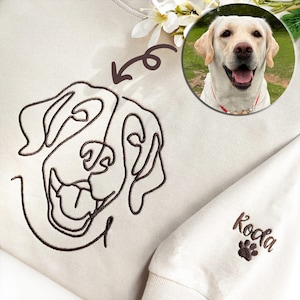 May include: Cream-colored sweatshirt featuring a brown line drawing of a dog's face. A circular photo of a smiling dog is in the upper right corner. The sleeve has the name "Koda" and a paw print embroidered in brown.