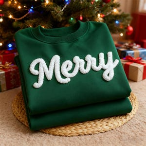 May include: A folded, dark green sweatshirt with the word "Merry" in white, textured lettering. The sweatshirt is displayed on a woven mat, with a Christmas tree and wrapped gifts in the background.