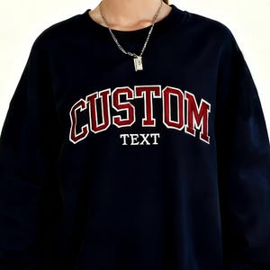 May include: Navy blue sweatshirt with the word "CUSTOM" in red and white collegiate lettering. Below the word "CUSTOM" is the word "TEXT" in white block letters. A silver necklace is visible.