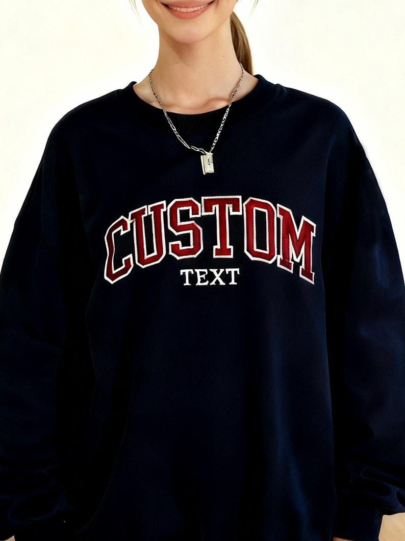 May include: Navy blue sweatshirt with the word "CUSTOM" in red and white collegiate lettering. Below the word "CUSTOM" is the word "TEXT" in white block letters. A silver necklace is visible.