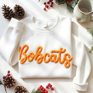 Può includere: Una felpa bianca con la scritta "Bobcats" in lettere arancioni strutturate. La felpa ha maniche lunghe e girocollo. L'immagine è decorata con pigne, bacche e una tazza.