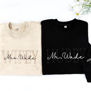 May include: Two matching sweatshirts, one cream and one black. The cream sweatshirt says "WIFEY Mrs. Wade" in embroidered lettering. The black sweatshirt says "HUBBY Mr. Wade" with the date "10.03.2025" and rings embroidered on the sleeve.