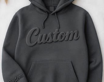 May include: A charcoal gray hoodie with the word "Custom" embossed across the chest and on the left sleeve. The hoodie features a drawstring hood and a front kangaroo pocket. The fabric appears soft and comfortable.