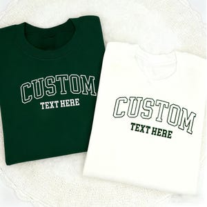 May include: Two sweatshirts, one dark green and one white, are displayed. Each has the word "CUSTOM" arched across the chest in a collegiate font, with "TEXT HERE" below. The text is in white on the green sweatshirt and green on the white one.