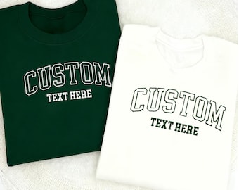 May include: Two crew neck sweatshirts, one dark green and one white. Both sweatshirts feature the word "CUSTOM" in an arched, collegiate-style font, with "TEXT HERE" underneath. The text is in a contrasting colour.