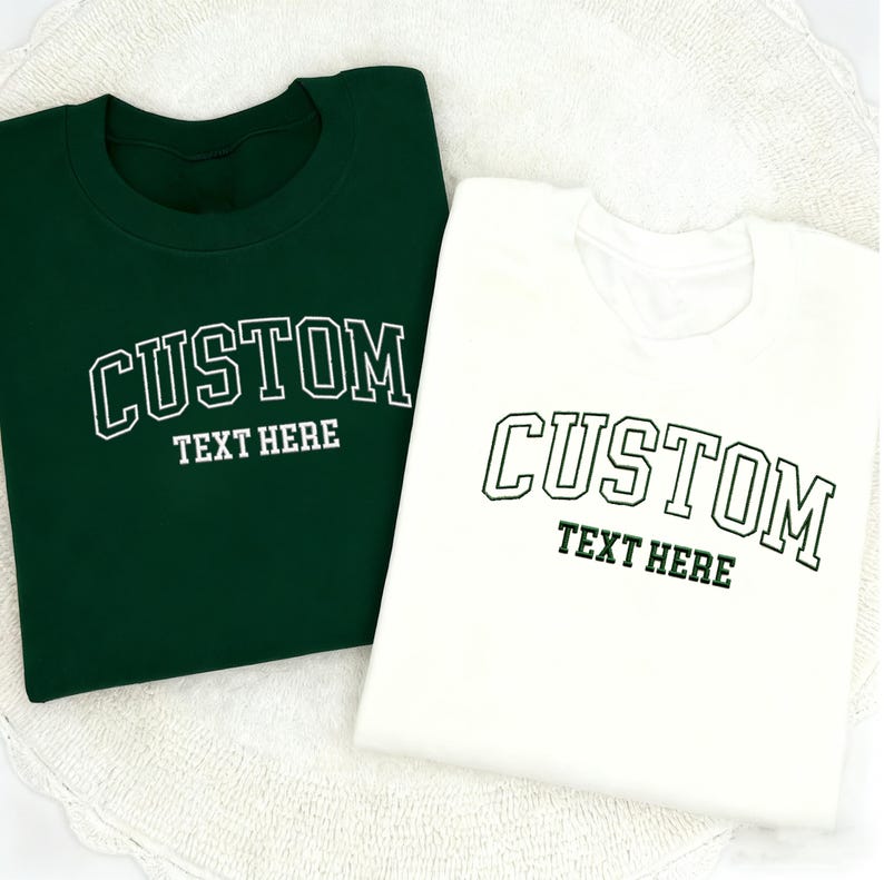 May include: Two sweatshirts, one dark green and one white, are displayed. Each has the word "CUSTOM" arched across the chest in a collegiate font, with "TEXT HERE" below. The text is in white on the green sweatshirt and green on the white one.