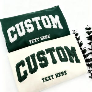 Puede incluir: Se muestran dos sudaderas, una verde oscuro y otra crema. Ambas presentan la palabra "CUSTOM" en letras bordadas grandes y en negrita. Debajo, el texto dice "TEXT HERE". Las sudaderas están sobre un fondo blanco.
