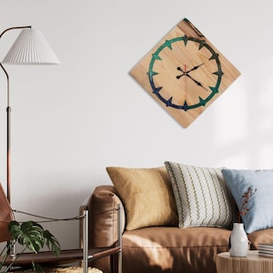 May include: A square wooden wall clock with a light wood grain finish. The clock face features a dark blue and green circular design with black clock hands and a red second hand. The clock is mounted on a white wall.