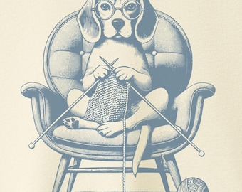 Puede incluir: Camiseta color crema con una ilustración azul de un beagle con gafas tejiendo en una silla. El perro sostiene agujas de tejer e hilo, con una madeja de lana en el suelo. La camiseta tiene cuello redondo y mangas cortas.