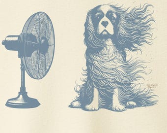 May include: Cream-colored t-shirt featuring a blue illustration of a fan and a Cavalier King Charles Spaniel with wind-blown fur. The dog is sitting, and the fan is to the left. The shirt has a crew neck and short sleeves.
