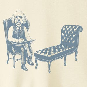 May include: Cream-colored t-shirt featuring a blue illustration of a dog in a suit sitting in a chair, holding a notepad, with a chaise lounge next to it. The shirt has the text "TACO DOG" on the upper right.