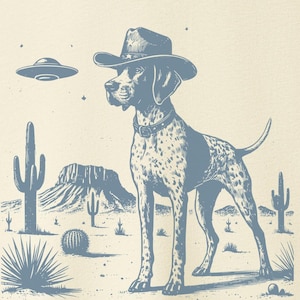 May include: Cream-colored t-shirt featuring a blue graphic of a dog wearing a cowboy hat in a desert landscape with a UFO. The dog is a pointer breed with spots. The desert scene includes cacti and a mesa.