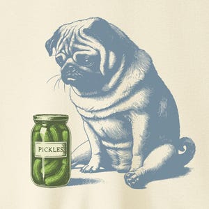 Op de afbeelding: Crèmekleurig T-shirt met een blauwe illustratie van een mopshond en een pot augurken. De pot is groen met het woord "PICKLES" erop gedrukt. De mopshond zit naast de pot en kijkt naar beneden.